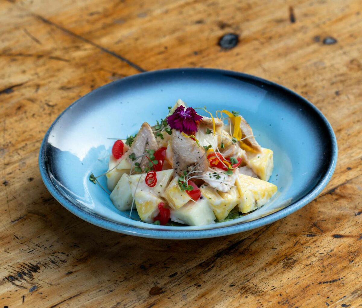 A vibrant tropical fruit and chicken salad served in a blue bowl on a rustic wooden table.