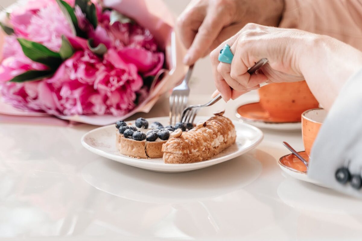 A delightful setup with hands enjoying a blueberry tart alongside a bouquet of pink flowers on a dining table.